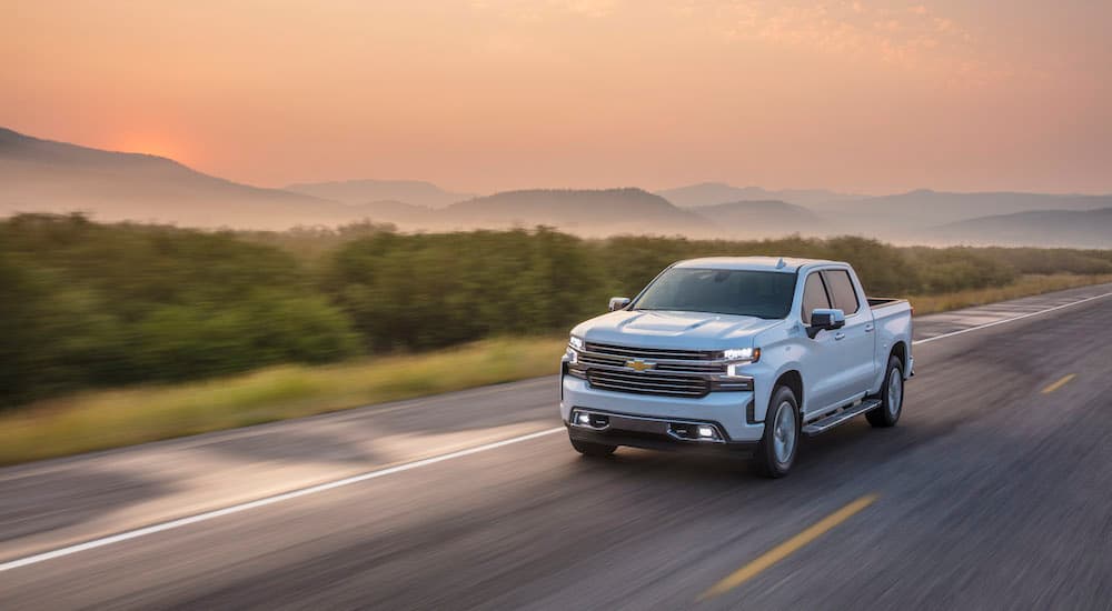 A popular vehicle in pre-owned luxury truck sales, a white 2021 Chevy Silverado High Country, is shown driving past distant mountains.